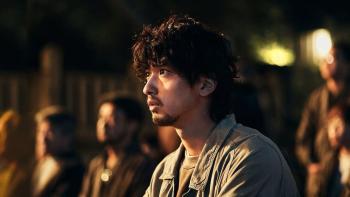 The Village (2023): A Japanese man center sits looking forward and left toward a stage performance