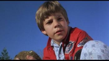 The Gate: Young boy in red jacket holding a rock, blue sky background.