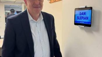 The Widower: Man in a suit standing beside a door labeled with a name on a digital sign.
