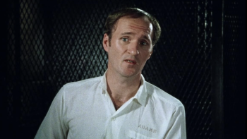 The Thin Blue Line: Man in white shirt stands by a dark textured background speaking to camera as part of an interview