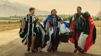 To the Max Movie: Three people in wingsuits walking on a road, smiling, with a mountainous backdrop.