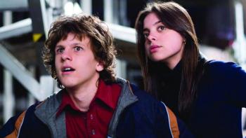 A close-up of Dyllan Christopher and Gia Mantegna: he is wearing a colored red shirt with a jacket, and she is wearing a black turtleneck.