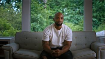 Untold: Shooting Guards movie: Ex-NBA player Javaris Crittendon in white shirt center sits on couch with green trees behind him through windows