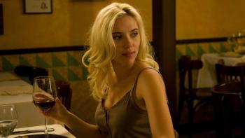 Scarlett Johansson sits at a restaurant table holding a glass of red wine, looking over her shoulder. 