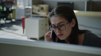 Still from Suspect/Victim: Investigative reporter Rachel de Leon, a White journalist, seated at a newsroom cubicle and looking downward at the desk wh