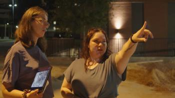 Still from Victim/Suspect: Investigative reporter Rachel de Leon, a White journalist (left) holding a tablet in her right hand, while listening to Emm