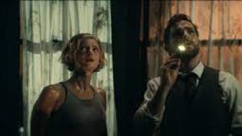 Vindicta Movie: A blonde woman and a White man in a dilapidated building shine a flashlight at something on the ceiling