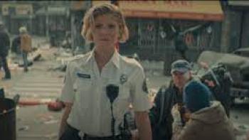 Vindicta Movie: A blonde female paramedic looks straight ahead on a dusty, chaotic street