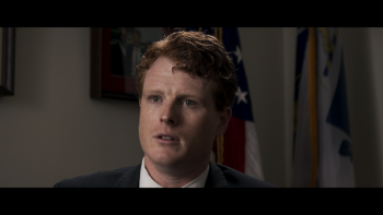 Still from Wake Up: Stories from the Frontlines of Suicide Prevention: Rep. Joe Kennedy III, a White U.S. Congressman, faces the camera and is seated for a firsthand interview, wearing a white shirt, dark-colored jacket, and reddish-brown colored hair