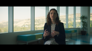 Still from Wake Up: Stories from the Frontlines of Suicide Prevention: Morissa Sobelson of the Harvard Injury Control Research Center facing the camera during a firsthand interview wearing a dark-colored jacket, diagonally-striped black and white dress, and shoulder-length brunette slightly wavy hair