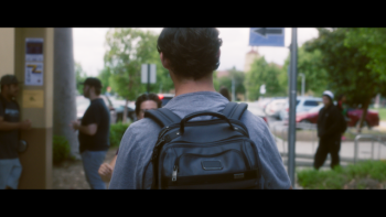Still from Wake Up: Stories from the Frontlines of Suicide Prevention: Reenactment of actor portraying Kellar Zibilich walking on college campus with back to the camera, wearing a gray-colored T-shirt, black backpack, and dark hair