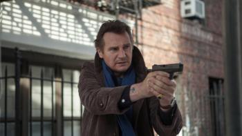 A Walk Among the Tombstones: Man in brown coat and blue scarf pointing gun with serious expression on urban street.