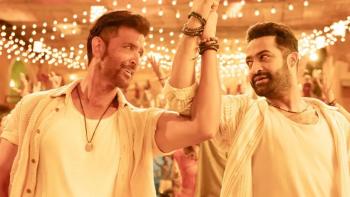 War 2: Two men in white clothing celebrating with raised clasped hands under string lights