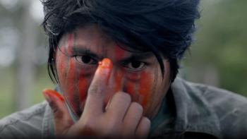 We Are Guardians: An indigenous man uses his pointer finger to paint his face with bright orange red paint