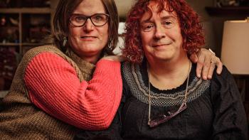 Still from We Live Here: The Midwest: Seated and facing the camera are Jenn Richmond (left) and Debb Richmond who are a White LGBTA+ couple from Minnesota, wearing casual attire