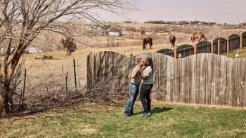 Still from We Live Here: The Midwest: Embracing each other while standing on farmland in Nebraska are Mario Foreman-Powell (left) and Monte Foreman-Powell, a Black and married LGBTA+ couple, wearing casual attire 