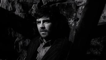 Whistle Down the Wind: A White middle-aged bearded man sits on straw against a wall in a barn while wearing a suit