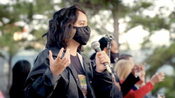 Still from Who We Become: With a microphone in the left hand, Filipino community organizer Jenah Maravilla addresses a diverse group of people at a stop Asian hate crime vigil and rally in Houston, Texas, wearing a black windbreaker jacket, dark-colored graphic top, and a black face mask
