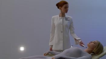 Wifelike: A woman in a white coat holds an implement to Elena Kampouris' neck as she lays down on a bed in front of her.