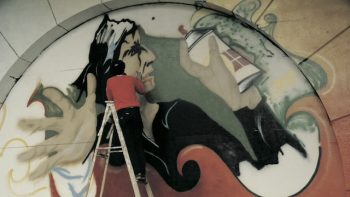 Still from Wild Style: Raymond a.k.a. Zero ("Lee" George Quiñones) with his back facing the camera while graffiti painting a New York City amphitheate