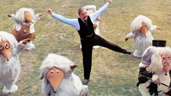 Wombling Free: An older White man in a waistcoat and tie dances among furry Wombles