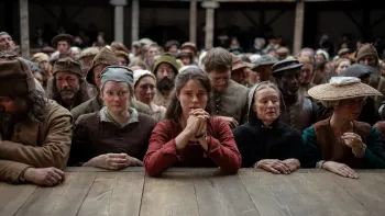 Hamnet: A young woman in red with clasped hands stands among a crowd in period costume at a wooden railing