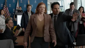 A hispanic man puts his hand up guiding a redheaded professional looking woman through a crowd of reporters at a press conference.