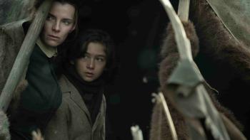 American Primeval TV show screenshot 1: Betty Gilpin and Preston Mota hide in a tent. Gilpin wears a black dress, and Mota wears a grey coat.