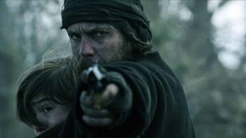 American Primeval TV show screenshot 2: Taylor Kitsch holds Preston Mota to his chest as he aims a gun. He wears a black coat, fingerless gloves.