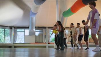 A group of dancers are led by Mor Hamami in a studio. She wears a white tank top and dark sweatpants.