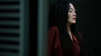 A Korean woman with wet hair and some visible wounds under her red blouse, stands with her shoulders high and her back against the wall