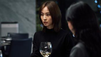 A Korean woman with shoulder-length brown hair sits in a darkened restaurant with a glass of white wine in front of her looking at someone off camera 