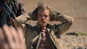Atomic TV show screenshot 1: Alfie Allen kneels holds his arms above his head. He wears a red shirt and a tan jacket. A man points a gun at his back.