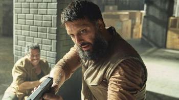 Atomic TV show screenshot 2: Shazad Latif points a handgun. He wears an orange shirt and a tan vest. A man leans on a brick pillar behind him.