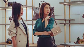 At the Moment TV show screenshot 3: Two women stand in a boutique, the woman on the right models a tan handbag.
