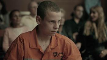Guy Manster sits in a court room. He wears an orange button-up shirt. A crowd sits in the background.