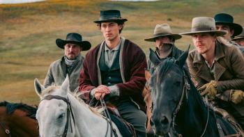 A group of five men dressed in Western-style gear sit atop horses standing close together. Behind them, a rolling hill and a bit of the sky.