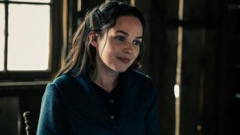 A smiling woman with blue eyes and long, dark hair is pictured from the chest up. She is wearing a navy blue dress with long sleeves and is sitting in a chair in front of a window. Some sunlight peeks through the window and through slats in the wooden walls.