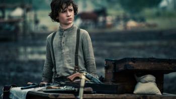 A young boy with brown hair is pictured standing outside, behind a wooden table covered with various guns. He wears a grey shirt and suspenders, and looks offscreen with a furrowed brow.