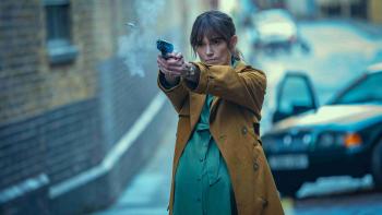 Black Doves TV show screenshot 3: Keira Knightly fires a gun while standing in the street. A baby bump pokes from a green dress and brown coat.