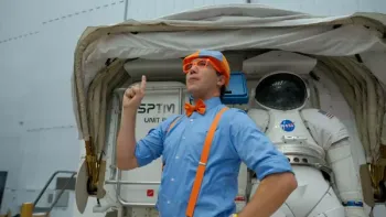 Blippi's NASA Astronaut Adventure TV: Blippi looks thoughtfully to the left holding one finger up. Behind him is an astronaut suit.