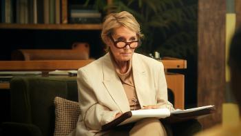 Blue Therapy TV show screenshot: A white woman in her 50s with glasses sits in an office wearing a blazer with a notepad on her lap