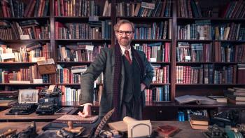 Bookish TV show screenshot 1 :Mark Gatiss stands in front of a cluttered bookshelf. He wears a green suit, red tie, white shirt, and a long scarf.