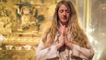 A white woman wearing white clothing sits in prayer position with eyes closed and cascading hair. She is in front of golden background.