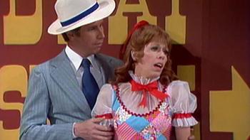 The Carol Burnett Show screenshot 1: Two actors from the Carol Burnett show appear in a skit. One is a man in a grey suit and white hat, the other is a redheaded woman in a colorful red, white, and blue dress.
