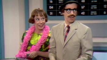 The Carol Burnett Show screenshot 3: Two actors from the Carol Burnett show appear in a skit. A redheaded woman at left holds a bag and is wearing a hot pink scarf. At right, actress Carol Burnett is dressed as a man with a black wig, mustache, and khaki suit.