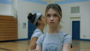 Chris, in a gym uniform and with arms crossed, looks serious, with another girl blurred in the background.