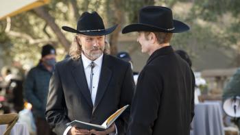 Two men in black suits and Stetson hats stand outdoors speaking with one another. One holds open a book.