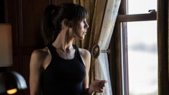 A brunette woman wearing a black tanktop stands near a window, gazing out.