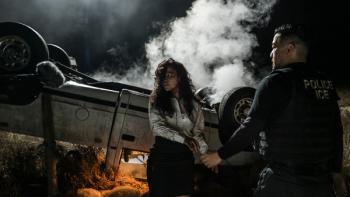 An overturned white truck is pictured with smoke coming out. A woman with curly black hair stands in front of the wreckage